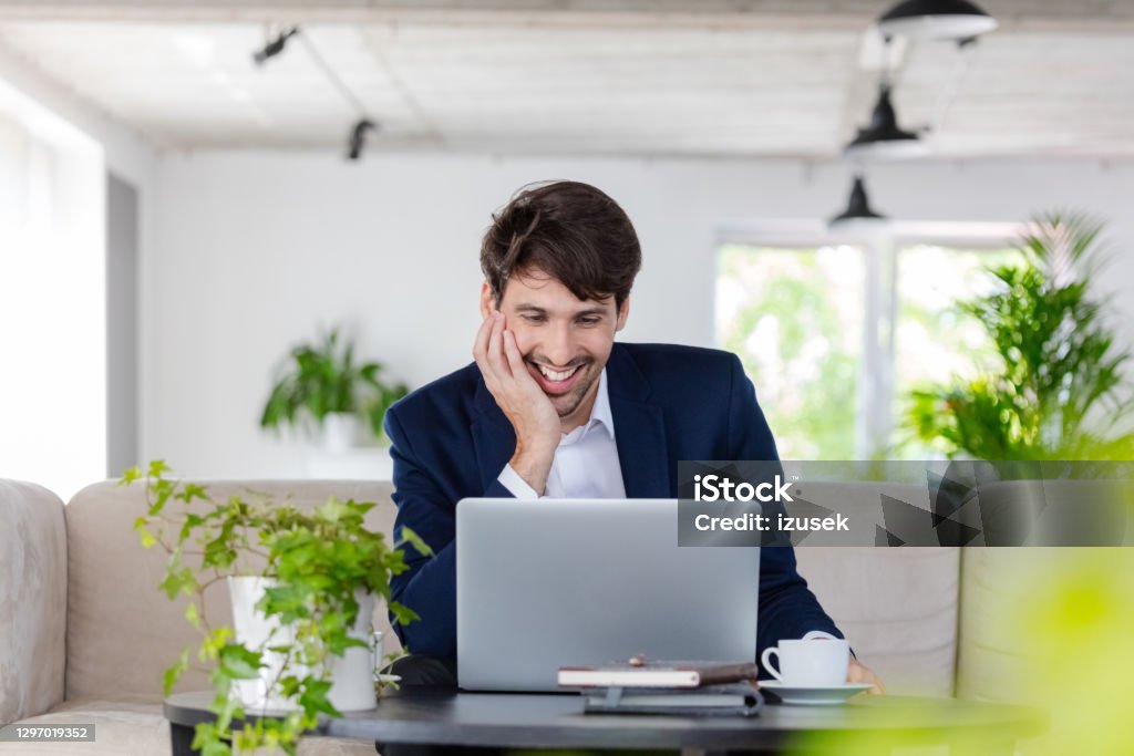 Businessman during video conference Cheerful mid adult man wearing navy blue jacket sitting on sofa in the creative workplace and using laptop during video call. 30-39 Years Stock Photo
