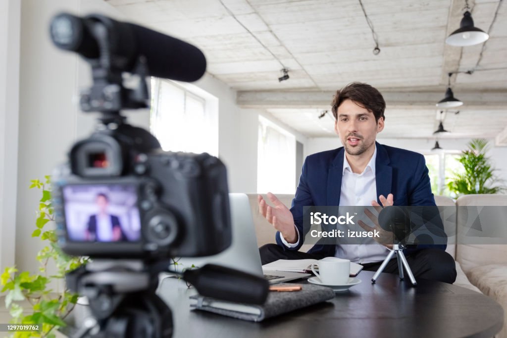 Businessman making a video blog in the office Mid adult businessmen wearing navy blue suit making a video blog. Vlogger recording content on digital camera in the creative office. 30-39 Years Stock Photo