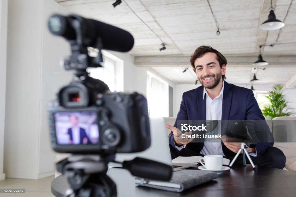 Cheerful businessman making a video blog in the office Mid adult businessmen wearing navy blue suit making a video blog. Vlogger recording content on digital camera in the creative office. Business Stock Photo