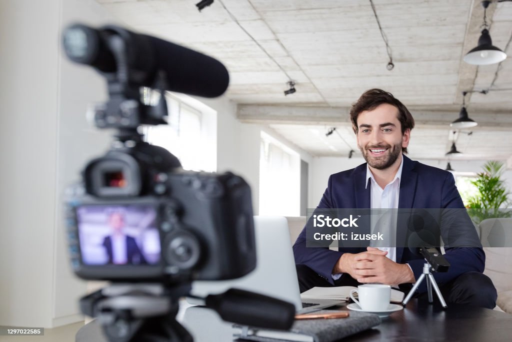 Cheerful businessman making a video blog in the office Mid adult businessmen wearing navy blue suit making a video blog. Vlogger recording content on digital camera in the creative office. 30-39 Years Stock Photo