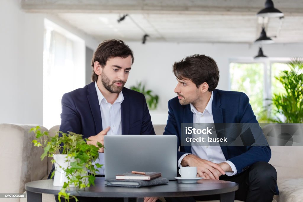 Shareholders discussing in an office Thoughtful mid adult businessmen wearing navy blue jackets and white shirts sitting on sofa in the green office and discussing over laptop. 30-39 Years Stock Photo