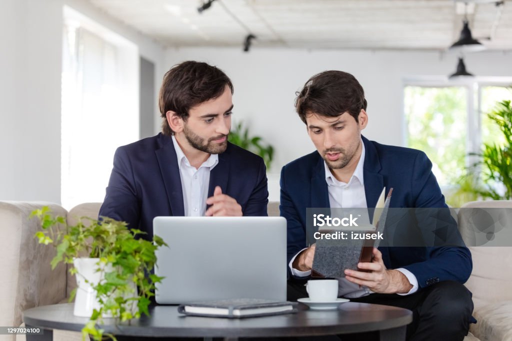 Two businessmen discussing in an office Thoughtful mid adult men wearing navy blue jackets and white shirts sitting on sofa in the green office and discussing over laptop, checking agenda. 30-39 Years Stock Photo