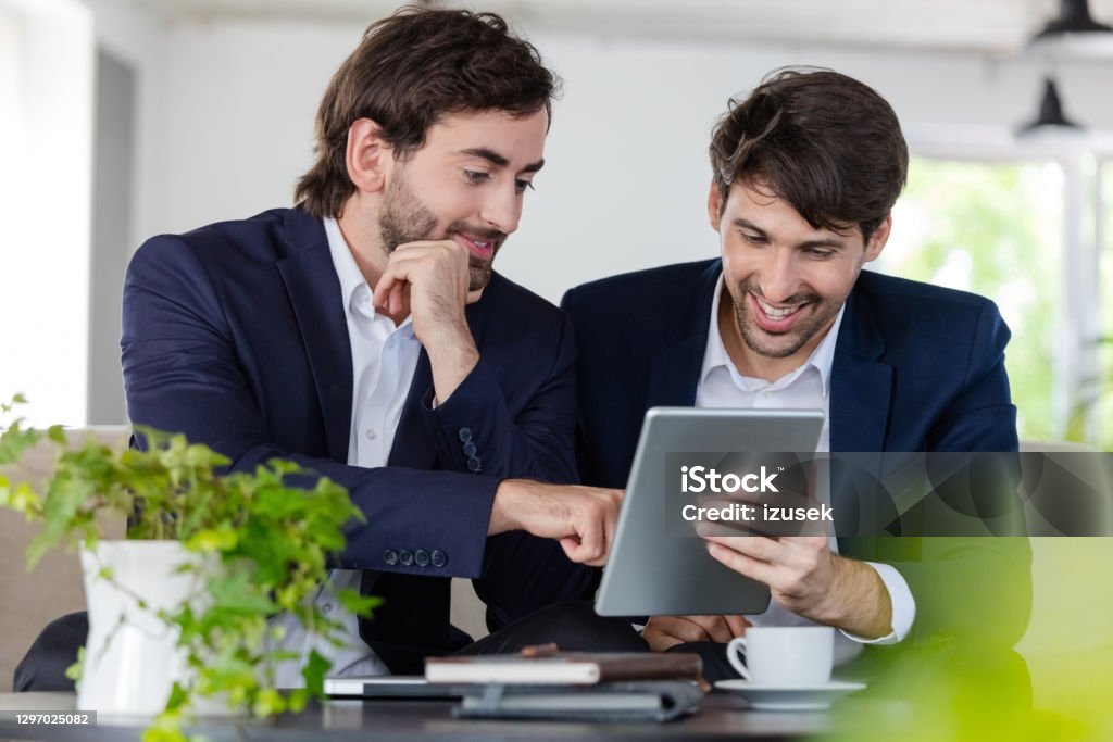 Two cheerful businessmen working together Cheerful mid adult men wearing navy blue jackets and white shirts sitting on sofa in the office and discussing over digital tablet. Shareholder Stock Photo