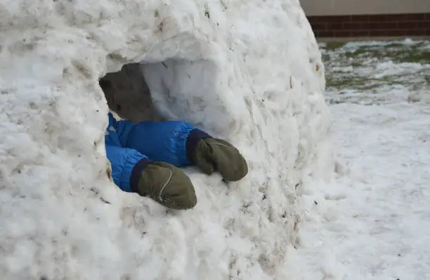 Photo of A small child in winter coveralls walks will build an igloo. little boy sticks his hands out of an ice cave, hole, house. He has gloves off his fingers and tries to get out of the freezing hole