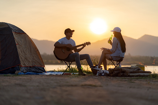 Young adult Asian couple playing guitar and drinking beer beside their tent campsite while sunset. Camping trip with dog outdoor activity campsite concept.