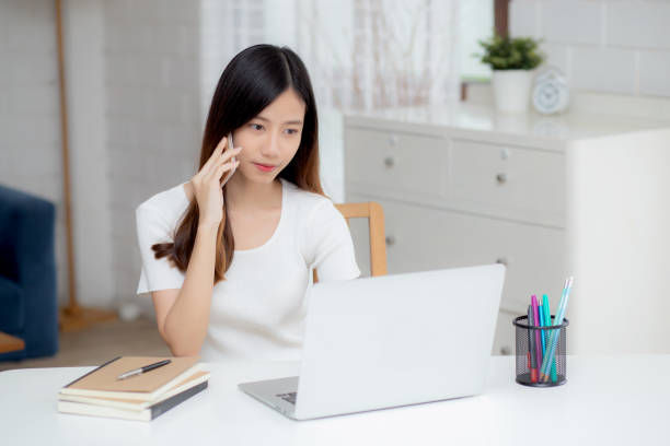 istock Young asian woman talking phone and work from home with laptop computer, freelance using notebook to internet online and speak on smartphone, domestic life, business and communication concept. 1297037730