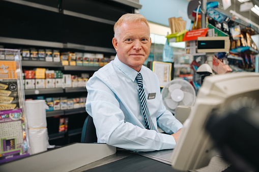 Portrait of a senior man working at super market checkout and looking at camera. Grocery store cashier at checkout counter.