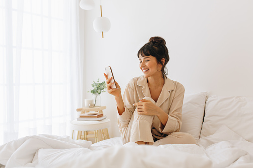Smiling woman sitting on bed and using mobile phone. Woman in night suit sitting on bed at home and relaxing looking at mobile phone.