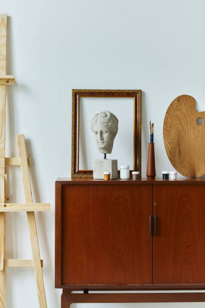 istock Stylish composition of artist workspace room with design retro teak commode, mock up poster frame, book, decoration and painting accessories. Template. 1297039774
