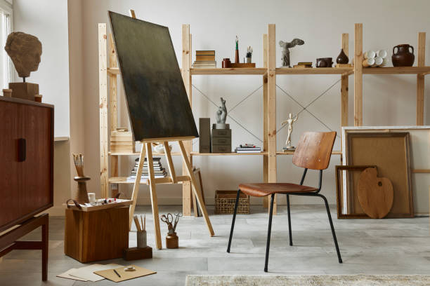 istock Unique artist workspace interior with stylish teak commode, wooden easel, bookcase, artworks, painting accessories, decoration and elegant personal stuff. Modern work room for artist. Template. 1297039775