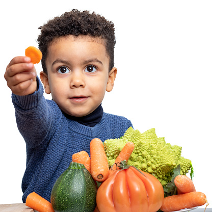 Close up portrait of cute afro american boy showing piece of carrot at table. Kid isolated on white background.