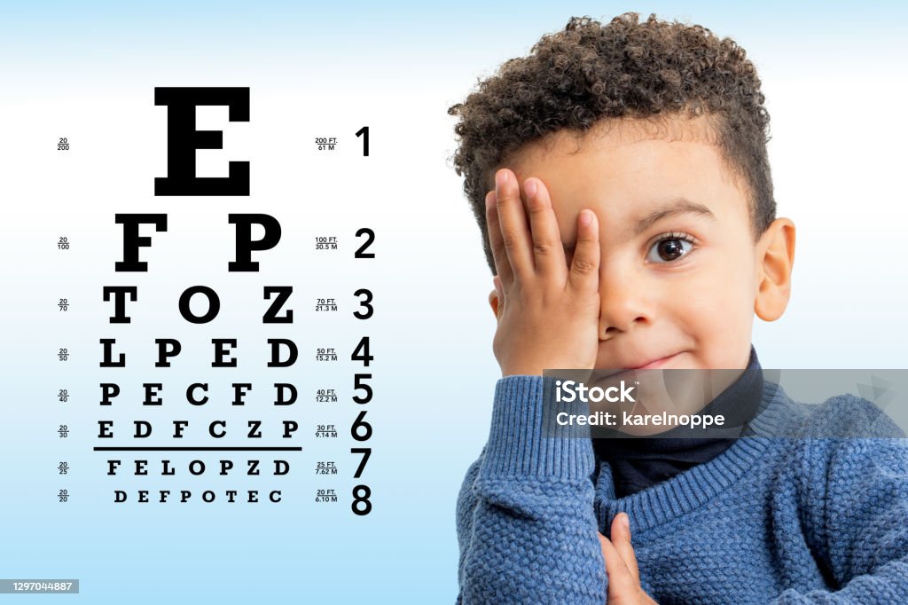 Cute afro american boy reviewing eyesight. Close up face shot of little Afro American boy testing vision. Kid with closing on eye with hand. Vision chart with block letters in background. Child Stock Photo