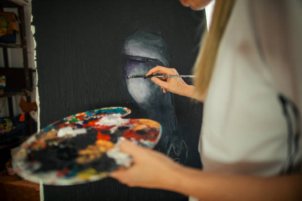 Beautiful woman artist painter standing near the easel in her atelier and working on her new project. She is holding painting brush and colorful palette. Focusing on her new painting with dark base color. She owns art and craft small business.