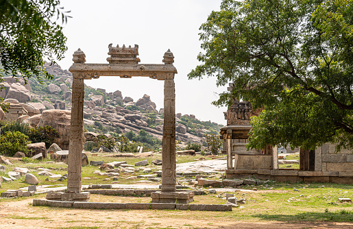 Hampi, Karnataka, India - November 5, 2013: Ruin of Ancient Temple. Closeup of beige stone sculpted free-standing portal with green hill full of brown stone boulders under silver sky. Green foliage around.