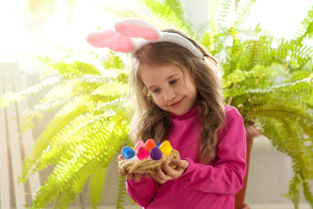 la petite fille mignonne 5-6 ans dans des oreilles de lapin admire un panier en osier avec les oeufs colorés de pâques sur le fond d’une fleur verte à la maison. enfance heureuse et concept de maison de pâques. copyspace - child caucasian little girls 3 4 years photos et images de collection