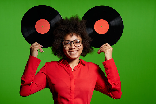 Funny African-American girl in red shirt holding vinyl disc as ears, over green background