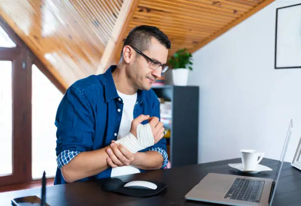 Businessman with bandaged wristis suffering from wrist joint pain at home office. Office syndrome hand pain by occupational disease.