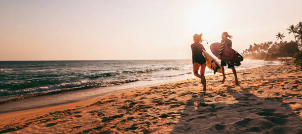 joven pareja hermosa caminando a lo largo de la playa de arena cerca del océano al atardecer con tablas de surf, actividades al aire libre y vacaciones deportivas - women sea cheerful surfing fotografías e imágenes de stock