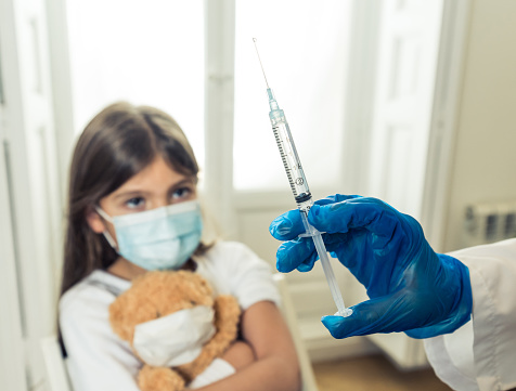 Nurse administering the coronavirus vaccine to a a young girl patient with face mask in Doctors clinic. Immunization, medical treatment and Covid-19 vaccination program after clinical trial in humans.