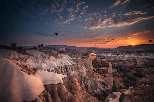 Spectacular sunset landscape view of hot air balloon illuminated with fire in sky tour over amazing fairy chimneys rock forms on valleys and fields in early morning in Kapadokya, Göreme National Park, Turkiye. Dreamy nature background.