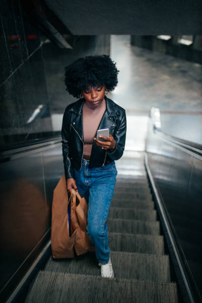 istock Afro woman using phone while she is on escalator and coming back from shopping 1297090231