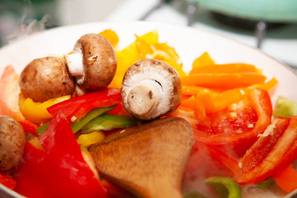 Sliced red, yellow, orange, and green bell peppers and whole portabella mushrooms in a frying pan with a wooden stirring spoon
