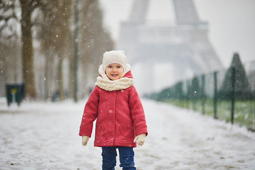 Adorable toddler girl near the Eiffel tower on a day with heavy snowfall in Paris, France. Happy child playing with snow. Winter activities for kids.