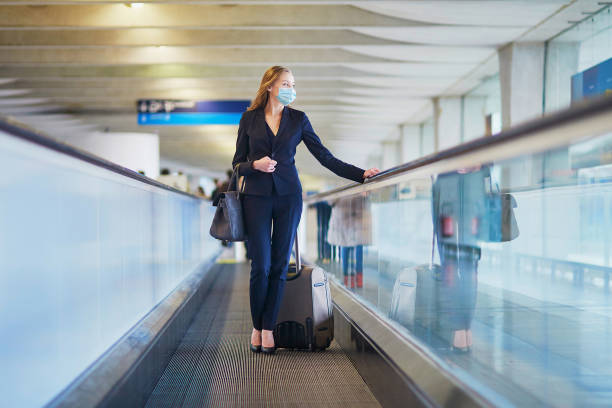 Business woman with hand luggage in international airport Young elegant business woman wearing protective face mask with hand luggage on travelator on international airport terminal. Cabin crew member with suitcase. Traveling during pandemic airport travelator stock pictures, royalty-free photos & images
