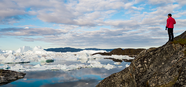 Travel adventure in Arctic landscape nature with icebergs - tourist person looking at view of Greenland icefjord - aerial photo. Man by ice and iceberg, Ilulissat Icefjord.