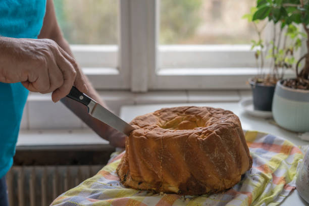 Men, visible just hand holding knife and cutting freshly baked Slovenian potica in domestic kitchen. He stands at the window.