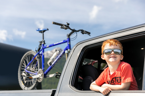 Young cute redhead child leaning on the car window and contemplating the view while on road trip. He is smiling and wearing sun glasses. He is going on a bicycle ride with his family.