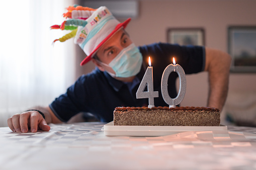 Mid adult man wearing a silly hat and a protective mask when blowing candles on his birthday cake