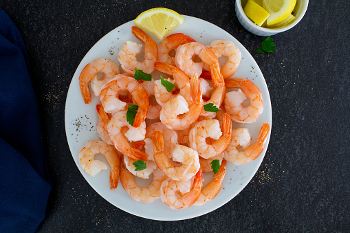 Freshly cooked shrimp served on a white plate with lemon and parsley.
Shot from above on a dark slate background. 
Close up.