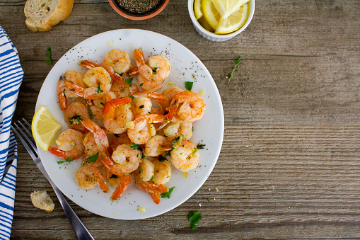 Panfried shrimp with garlic and herbs served on a white plate with lemon and fresh crusty bread.\nShot from above on a rustic wooden table. \nCopy space to the right of the image. \nFlat lay.