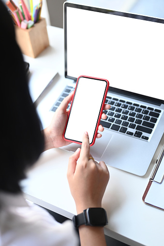 Mockup image of a woman holding smart phone with blank desktop screen while sitting in front of her laptop.