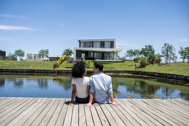 istock Couple sitting together on dock looking at house 1297128983