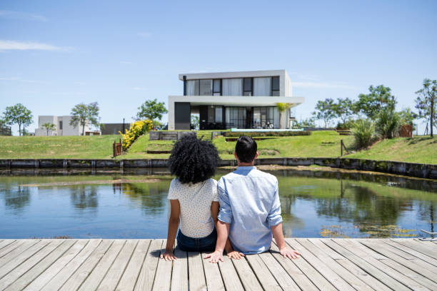 istock Couple sitting together on dock looking at house 1297129013