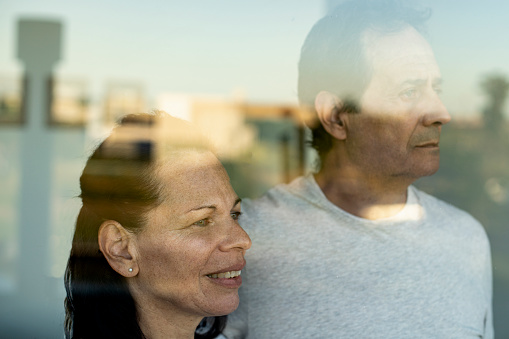 Thoughtful mature couple looking through window while standing at home