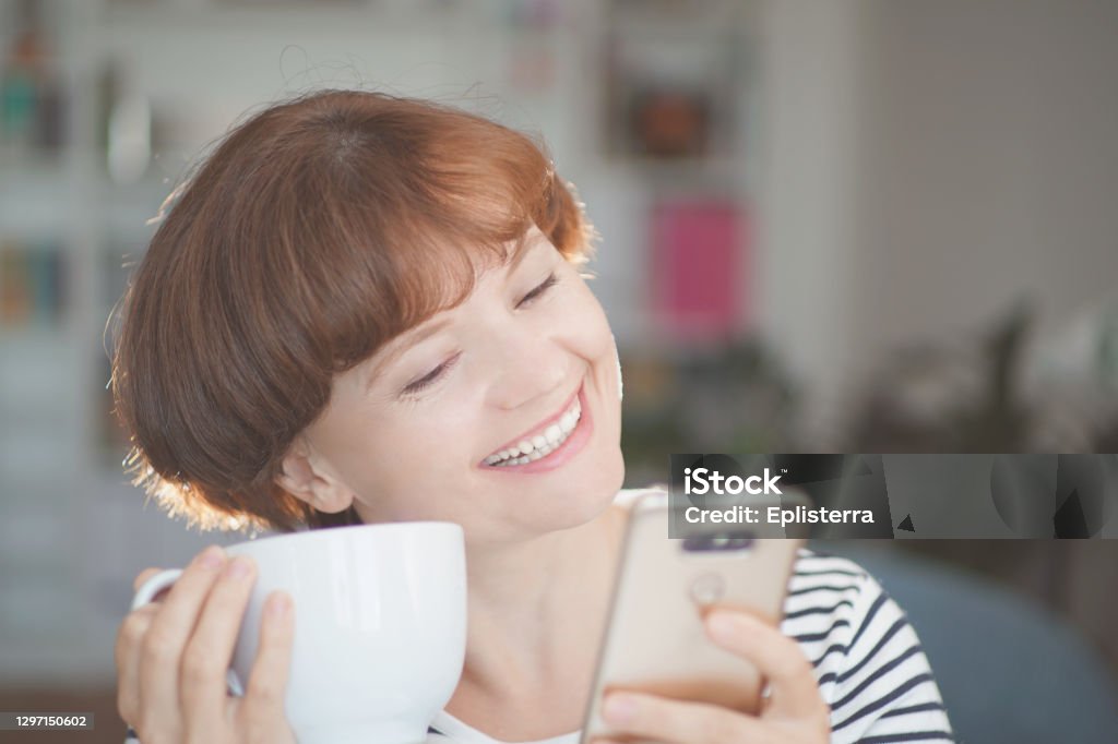 Young beautiful woman holding coffee cup, looking at smartphone and smiling. woman happy girl with cellphone. Home office and social media distance. Computer Network Stock Photo