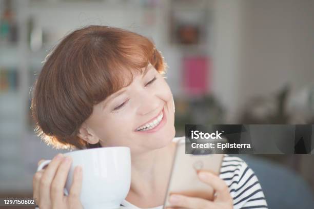 Young Beautiful Woman Holding Coffee Cup Looking At Smartphone And Smiling Woman Happy Girl With Cellphone Home Office And Social Media Distance Stock Photo - Download Image Now