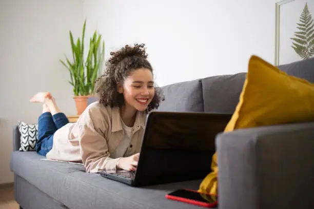 Photo of happy mixed rase girl is lying on couch and looking at at computer screen laptop. woman talks over Internet. Video Conference or e-learning at home.