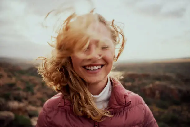Portrait of young smiling woman face partially covered with flying hair standing at mountain - carefree woman