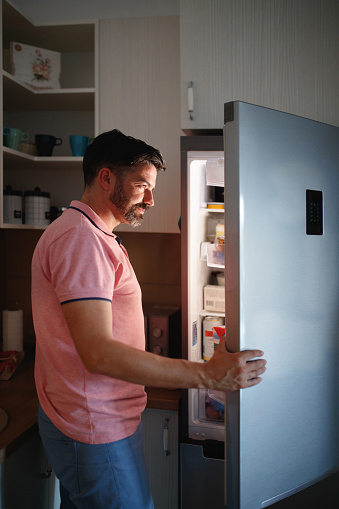 Man  in pink t-shirt looking inside the refrigerator and choosing groceries, his face is lit by refrigerator light