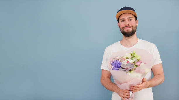 Delivery man with bouquet flowers Delivery man in a cap and white t-shirt with beautiful bouquet on a blue background. Flowers delivery concept. Banner with man with flowers man flower stock pictures, royalty-free photos & images