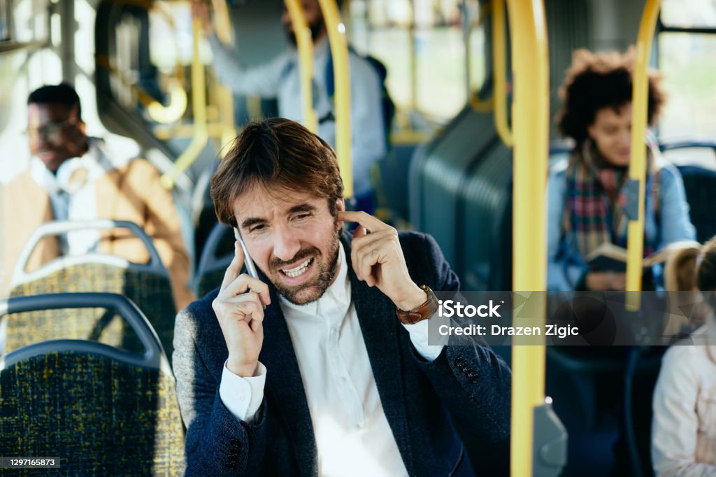 Speak louder please, I'm in a bus! Mid adult businessman talking on the phone and holding finger in ear while commuting by public transport. Noise Stock Photo