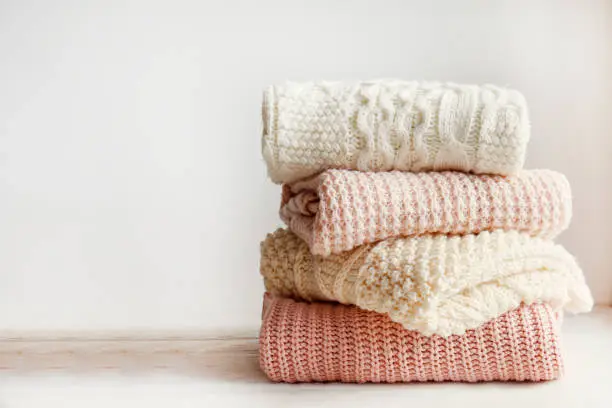 Stack of clean freshly laundered, neatly folded women's clothes on wooden table. Pile of shirts and sweaters on the table, white wall background. Copy space, close up, top view.