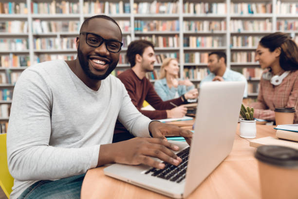 Group of ethnic multicultural students in library. Black guy on laptop.