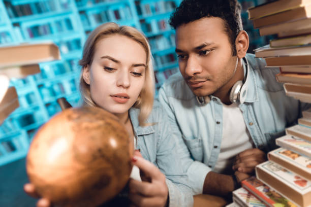 istock Ethnic indian mixed race guy and white girl surrounded by books in library. Students are using globe. 1297168873