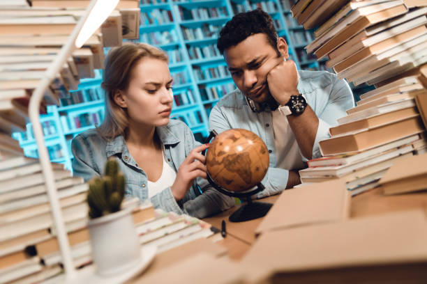 istock Ethnic indian mixed race guy and white girl surrounded by books in library. Students are using globe. 1297168883