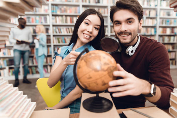 istock Ethnic asian girl and white guy surrounded by books in library. Students are using globe. 1297169530
